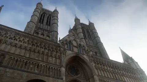The picture has been taken from the ground, and looks up at the cathedral. It is a gothic design, with a big archway in the centre. There are two steeples either side of the archway which extend into the sky. 