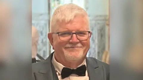 PSNI John Atkinson smiling. He is wearing a grey suit jacket, white shirt, black bow tie and black framed glasses.