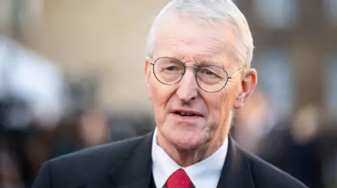 James Manning/PA Wire Hilary Benn, a man with short, grey hair, looks past the camera with a serious expression. He is wearing metal-framed glasses, a black suit jacket, a white shirt and a red tie. The background is blurred. 