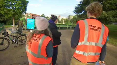 A group of three people in a park setting, all three have their backs to the camera. Two of them are wearing orange hi-viz vests which have Toxic Air Officer printed on the back