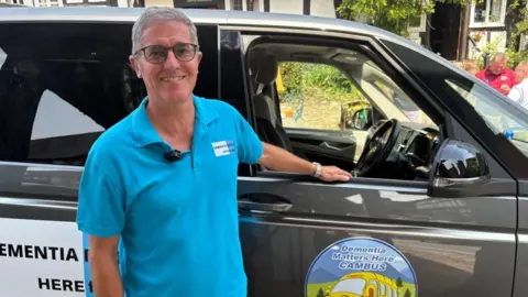 A man in a blue polo shirt, with dark grey hair and glasses, stands next to a gun-metal grey people carrier with a cartoon logo on the side. Behind the vehicle you can see a tudor style black and white building.