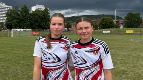 Two girls with brown hair in Crusaders FC away shirts (white with red and black markings) stand on a football pitch. High rise flats can be seen in the background, the sky has dark rainclouds.