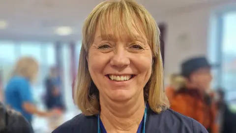 Helen Smith, with shoulder length blond hair wearing her nurse outfit at a support group with another health care worker and patient blurred in the background. She is smiling.