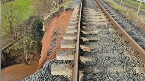 A picture of a railway track, where the embankment on the left-hand-side has fallen away in a landslip. A river can be seen below this.