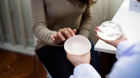 Woman sitting on a chair touching breast implants being held out someone else