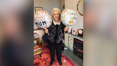 Kirstie Lumley A woman in her eighties is standing in her living room holding two helium balloons.  One says "Blooming Marvellous Mum" and the other says "Wonderful Grandma".