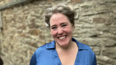 A woman looking at the camera and smiling. She has short hair. She is wearing a blue top. There is a blurred brick wall behind her.