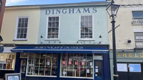 The shop front of Dinghams. It is a terraced building painted mint green, with the word Dinghams painted in navy capital letters just below the eaves. There is a navy shop awning, and the word 'sale' in the window. The shop window is full of items for sale.