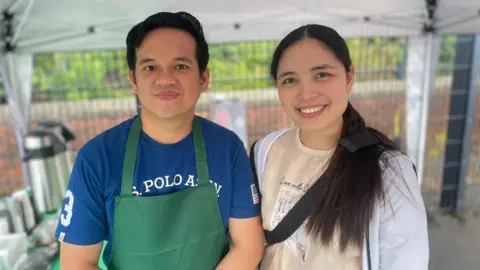 A man and woman stand side by side. The woman has long brown hair and wears a white shirt with a sand coloured top underneath. The man who stands to the woman's right wears a blue t-shirt with a green apron on over it, he has short brown hair.