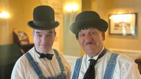 Two men dressed in black bowler hats, white shirts, blue denim dungarees and black ties smile at the camera.