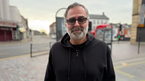 A man wearing a black hoodie and sunglasses stands in a street.