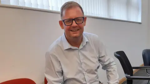 LDRS A man in a pale blue shirt and tan trousers sits on a chair and smiles. He has short brown hair and dark-framed glasses. A white wall and window can be seen behind him.