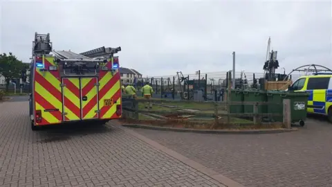 A fire engine and a police van are parked outside a play park named 'Shiver Me Timbers' in Maryport. Firefighters are waiting outside the fencing of the play park.