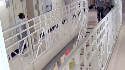 West Yorkshire Police A prison corridor with white railings and cells around the edge. There is a man walking down the corridor in a prison issue grey tracksuit. 