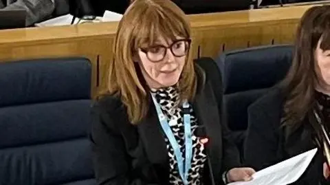 TraffordCouncil Councillor Jo Harding delivers her budget speech in Trafford's council chamber. She is holding on to a  bundle of papers, stood up above some blue chairs. 