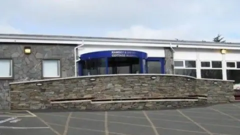 The front of the Ramsey hospital, which has a curved stone wall in front of the entrance of a large blue revolving door.