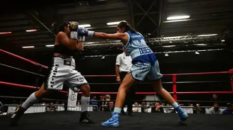 Fightzone Two women box in a professional fight. A referee and judges can be seen in the background.