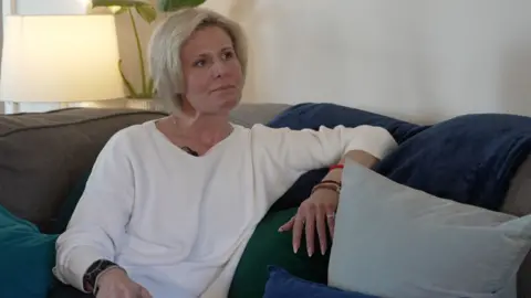 A woman with cropped blonde hair is wearing a long-sleeved white T-shirt. She is sitting on a dark-coloured sofa.