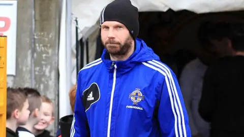 Irish Football Association/PA Wire Alan Crooks in blue IFA jacket, with a black beanie hat. He is looking at the camera, with some boys in the dugout behind him.