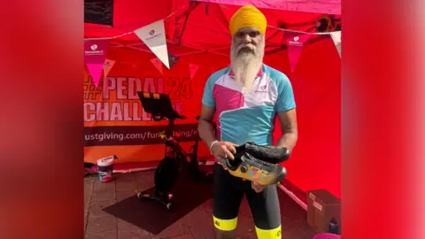 Manny Singh Kang A man in a white, pink and blue Dementia UK top on, and black and yellow cycling shorts. He has a yellow head scarf on, and has long white facial hair. He is holding a pair of trainers and is standing in front of a red tent with a bike machine in it.