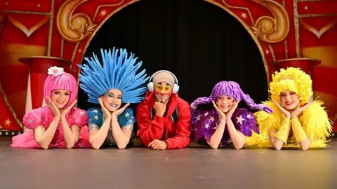 Andrew Payne Five people dressed in colourful circus outfits lie down on a stage with their heads in their hands.
