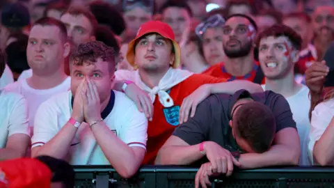 PA Media Men in red and white England shirts with a disappointed look on their face as England lose the Euro 2024 final to Spain. One man in a grey T shirt rests his head on his arm. 