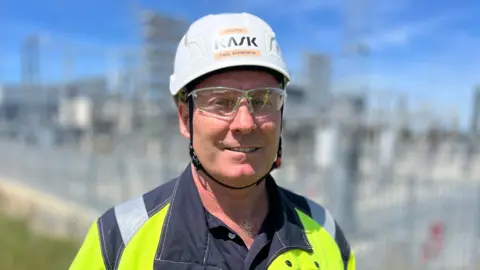 Martin Giles/BBC Paul Ashworth, a man standing at the 49-acre site of the onshore converter substation in Swardeston in Norfolk. He is looking directly at the camera and is smiling. He is wearing a black T-shirt with a fluorescent yellow hi-vis jacket over the top, a white hard hat and a pair of PPE glasses.