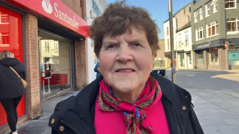 Margaret Sinclair has brown hair, worn short and a faint smile. She has a bright pink jumper on and a silk scarf around her neck. She wears a navy jacket 