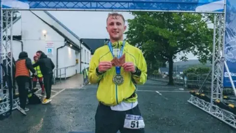 Callum Laird Callum Laird standing at the finish line of a marathon, proudly wearing a medal around his neck. He is wearing a yellow jacket and has a marathon number tag on his shorts. 