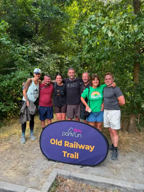 Josh Smith Seven people stood behind a parkrun-branded purple and orange sign which reads: Old Railway Trail. They have their arms around each other and are smiling and looking at the camera. Behind them are trees and bushes.