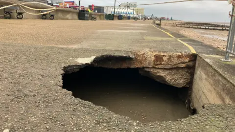 A sinkhole of around two square metres is shown filled in with concrete on a seaside promenade, with a concrete walkway and wooden benches shown in the background next to the beach and sea defences to the right of the image.