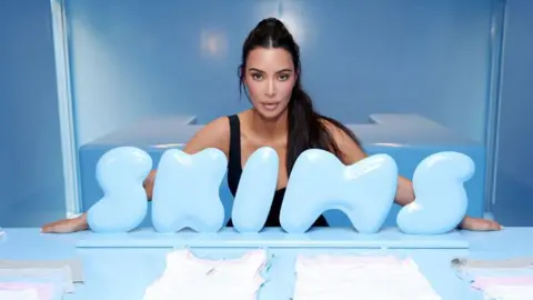 A empreendedora da Getty Images, Kim Kardashian, posa para uma foto posicionada atrás de um logotipo azul-celeste da Skims apoiado na lateral de uma mesa azul com as roupas de sua empresa.