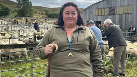 Shepherdess Cara Lowe is standing outdoors at the Lairg lamb sales. She has long brown hair and is wearing a light green fleece. She is holding a shepherd's crook and has a whistle to instructing sheepdogs hanging from a lanyard around her neck.