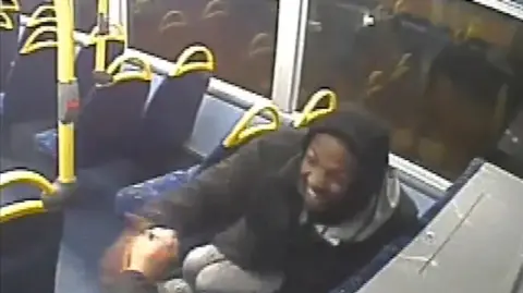 Metropolitan Police Dino Donaldson sits on a bus, with a black hood on, laughing. The hand of another person is also in the picture. There are empty bus seats in front of him. 