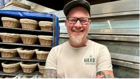 Linsey Smith/BBC Phil Clayton, a man wearing a hat and glasses and with tattoos on his arms, is smiling broadly as he stands in front of an industrial over. To the left of the image are racks full of baskets.