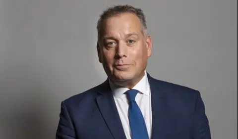 UK Parliament A head and shoulders image of David Warburton looking into the camera. He is wearing a suit and has short grey hair and 