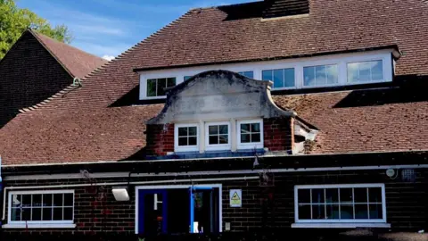BBC Outside of an infants school in Surrey. A red/brown brick building, which has faded letters in the middle, which say: 1905, BOYS SCHOOL'