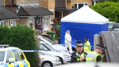 PA Media Police and a forensics officer at the scene outside the Hunt family home. The house is in a residential street with cars parked on driveways. There is a blue and white forensics tent up outside the house, which has been taped off.