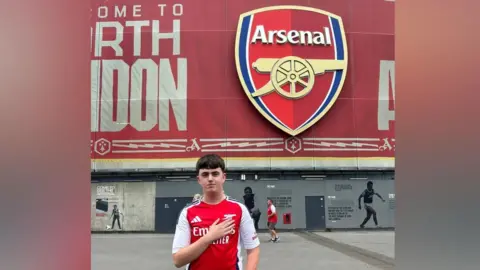 Alex had brown hair and is standing in front of the Emirates stadium which has a large red logo on it in the shape of a shield. It says Arsenal and has a large gold cannon on it. Alex is wearing a red and white Arsenal t-shirt and has his arm across his chest