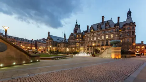 Getty Images Sheffield Town Hall photographed at dusk. A large stone-built building stands behind a paved area with a large fountain and grassy areas. 