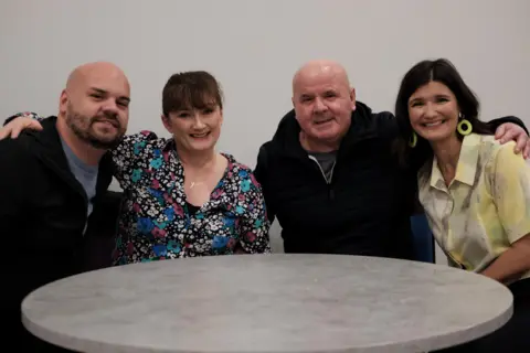 James snr (right) and James jnr (left) with River City actors Sally Howitt who plays Scarlett O'Hara and her on-screen daughter Kelly-Marie, played by Carmen Pieraccini. They are sitting around a round table and have their arms around each other.