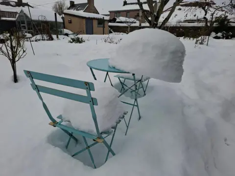 Ann Gray A garden covered in snow, with a large pile of snow slowly sliding off a table