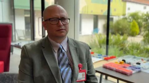 A bald man sits in a room with glazed wall and landscaped courtyard beyond. he is wearing a grey suit jacket, lilac shirt and blue and grey striped tie, and a red lanyard with his photo ID inside it. He is wearing black framed glasses and looking to the right of the camera.