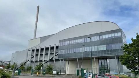 A large metal and glass incinerator with a tall grey chimney and cars and trees around it 