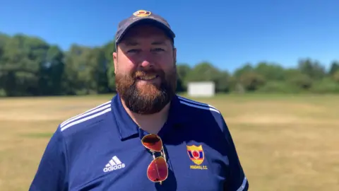 Maghull Cricket Club chairman Adam Lloyd smiles while stood on the club's cricket ground. He has a big beard and is wearing a cap and club t-shirt.