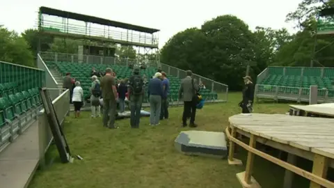 A temporary amphitheatre being constructed, with green fold-up seating and a number of people standing and working on the structures. Part of a wooden stage can be seen in the foreground