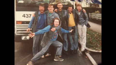Dave Callaghan Six men are stood in front of a bin lorry with the number 437. They are linking arms and smiling for the camera. One man is stood in front of the other five and spreading his arms wide.