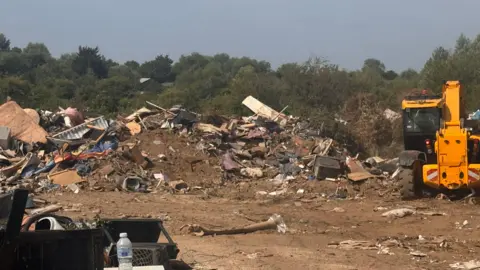 A site with a big pile of dumped waste. There is a yellow digger next to the pile and lots of trees in the background. 