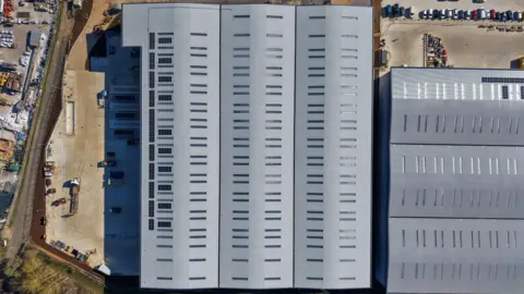 RECONOMY An aerial shot of a large grey warehouse style building taken from directly above the building. Solar panels can be seen on the roof. As well as some vehicles, bare soil can be seen on two sides of the building, showing construction at the site has not been completed.