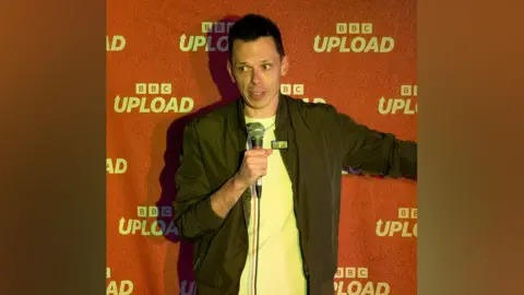 A man with dark hair wears a black jacket over a white t-shirt. He stands in front of a red backdrop which says "BBC Upload" on it.